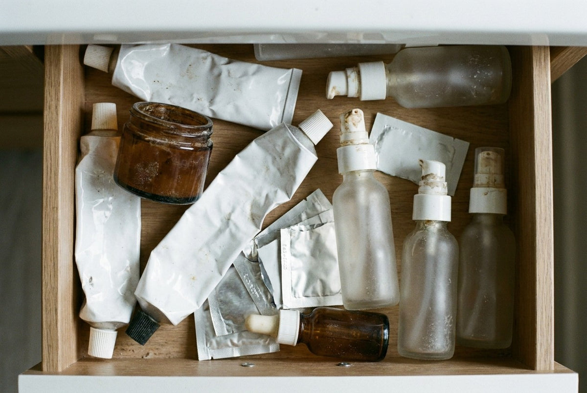 Overhead view into an open bathroom drawer holding eleven abandoned skincare products: creased tubes, a jar crusted at the rim, clogged pumps, torn sachets. All unbranded, all used, none finished.