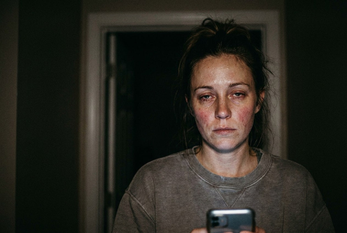 A woman in a dark hallway at three in the morning, her face lit only by the phone she is holding. Tired eyes, visible redness across the cheeks, no makeup.