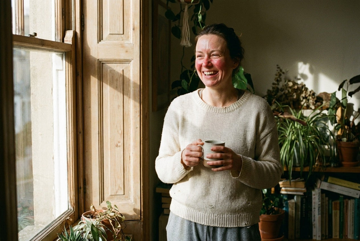 A woman in her late thirties laughing at something out of frame by a sunlit window, holding a mug. Her cheeks and nose are still clearly red, skin unretouched.